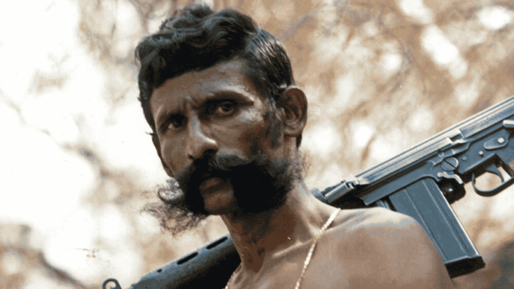 Close-up of poacher Veerappan with a thick mustache, bare-shouldered with a rifle slung over his shoulder, staring directly at the camera against a blurred outdoor background of trees