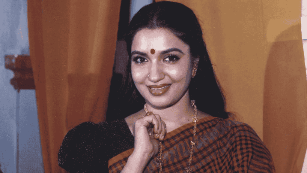Portrait of actress Sukanya smiling warmly, wearing a red-and-black checkered saree, gold jewelry, a bindi, and a nose ring, posing indoors in front of warm-colored curtains with one hand raised near her chin